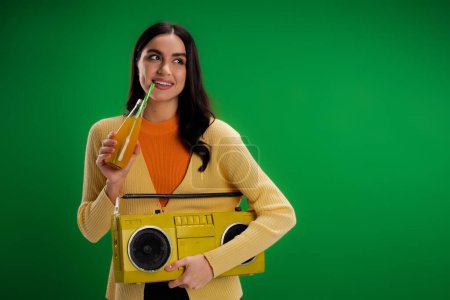 Foto de Smiling woman with boombox drinking soda and looking away isolated on green - Imagen libre de derechos
