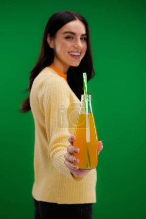 bottle with fresh lemonade in hand of brunette woman smiling on blurred background isolated on green