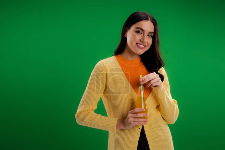 Foto de Young brunette woman with bottle of natural juice smiling at camera isolated on green - Imagen libre de derechos