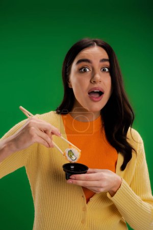 astonished woman holding sauce bowl and chopsticks with sushi roll while looking at camera isolated on green