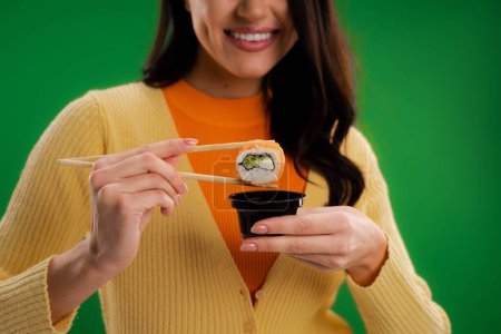 cropped view of smiling woman holding sushi roll near sauce bowl isolated on green
