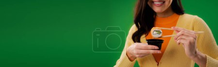 partial view of happy woman holding chopsticks with sushi roll near sauce bowl isolated on green, banner
