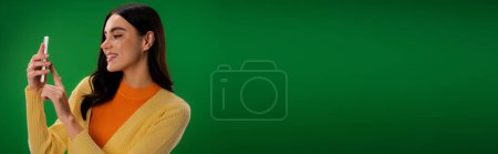 Téléchargez les photos : Pleased woman in orange turtleneck using smartphone isolated on green, banner - en image libre de droit