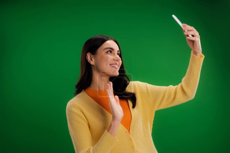 Foto de Happy and stylish woman waving hand during video call on smartphone isolated on green - Imagen libre de derechos