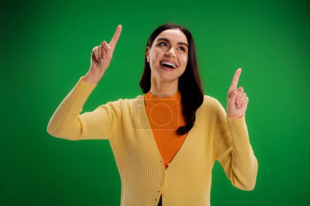 Foto de Excited woman in yellow jumper looking up and pointing with fingers isolated on green - Imagen libre de derechos