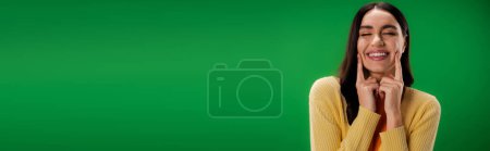 Photo for Young brunette woman with closed eyes touching cheeks and smiling isolated on green, banner - Royalty Free Image