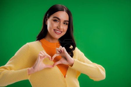 Téléchargez les photos : Pretty brunette woman showing heart sign with hands and smiling at camera isolated on green - en image libre de droit