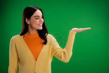 Foto de Smiling woman in orange turtleneck and yellow jumper pointing with hand isolated on green - Imagen libre de derechos