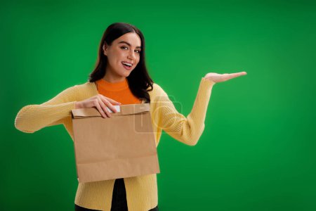pleased woman holding food package and pointing with hand isolated on green