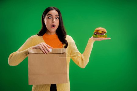 amazed woman with tasty burger and paper bag looking at camera isolated on green