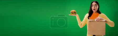 astonished woman looking at camera while holding paper bag and delicious burger isolated on green, banner