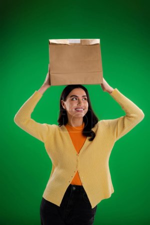 Foto de Cheerful young woman holding paper bag above head isolated on green - Imagen libre de derechos