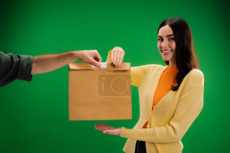 pretty brunette woman taking paper bag from delivery man and smiling at camera isolated on green