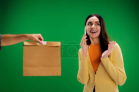 cheerful young woman calling on cellphone near man with food package isolated on green