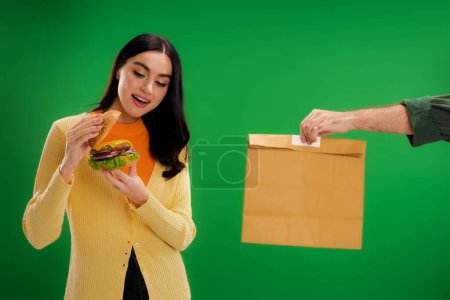 pleased woman looking at tasty burger near delivery man with paper bag isolated on green