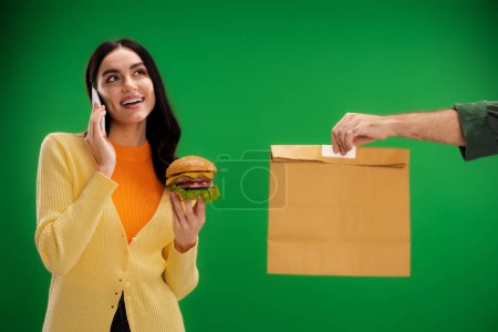 smiling woman holding burger and talking on smartphone near courier with paper bag isolated on green