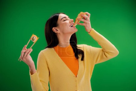 young brunette woman holding chopsticks with sushi roll while eating pizza isolated on green
