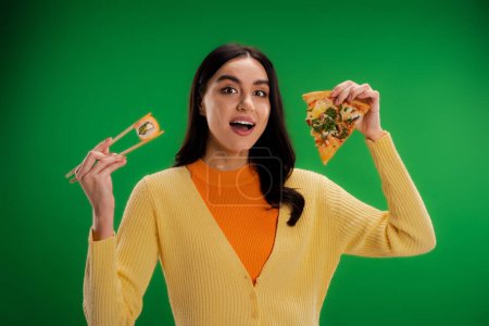 Photo for Amazed woman looking at camera while holding tasty pizza and fresh sushi roll isolated on green - Royalty Free Image