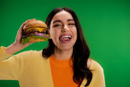 brunette woman holding tasty burger and sticking out tongue isolated on green
