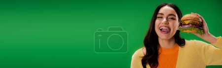 joyful woman with delicious burger sticking out tongue and looking at camera isolated on green, banner