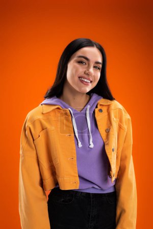 Téléchargez les photos : Young woman in purple hoodie and stylish jacket smiling at camera isolated on orange - en image libre de droit