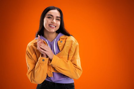 Téléchargez les photos : Happy brunette woman in trendy jacket and purple hoodie looking at camera isolated on orange - en image libre de droit