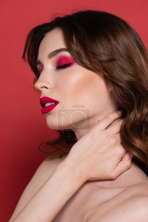 portrait of young woman with magenta color makeup touching neck isolated on pink 