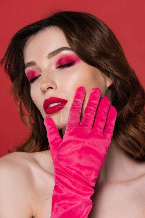 portrait of brunette woman with magenta color glove touching face isolated on pink 