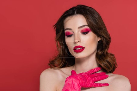 portrait of young woman with magenta color eye shadow touching chest with hand in glove isolated on pink 