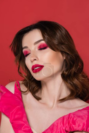 portrait of young woman with magenta color eye shadow embracing herself isolated on pink 