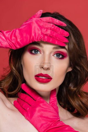 young brunette woman with magenta color eye makeup looking at camera while posing in gloves isolated on pink 