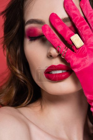 brunette young woman in magenta color glove with golden rings covering face isolated on pink 