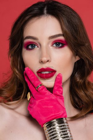 portrait of young woman in magenta color glove with golden rings and bracelet touching face isolated on pink 