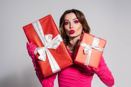 Téléchargez les photos : Brunette young woman in magenta color gloves holding presents while pouting lips isolated on grey - en image libre de droit