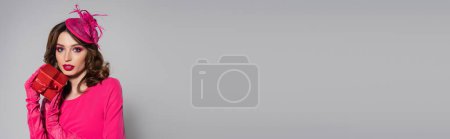 Téléchargez les photos : Brunette young woman in magenta color gloves and hat with feather holding present on grey, banner - en image libre de droit