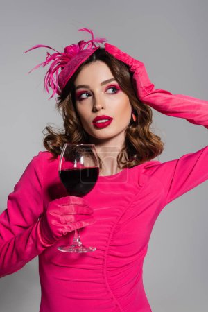brunette woman in magenta color gloves and elegant hat with feather holding glass of red wine isolated on grey 