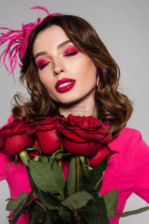 brunette young woman with magenta color makeup and hat with feather posing near red roses isolated on grey 