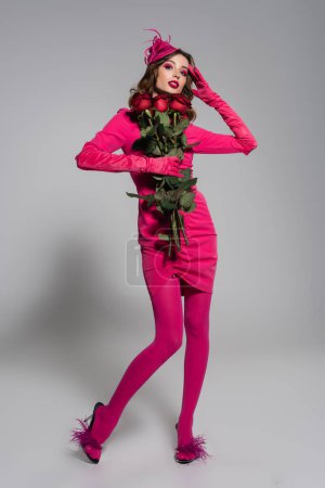 Foto de Full length of young woman in magenta color gloves and hat with feather holding red roses on grey - Imagen libre de derechos
