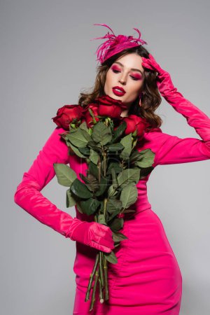 Téléchargez les photos : Brunette young woman in magenta color gloves and hat with feather looking at red roses isolated on grey - en image libre de droit
