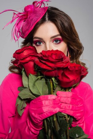 Photo for Brunette young woman in magenta color gloves and hat with feather covering face with red roses isolated on grey - Royalty Free Image