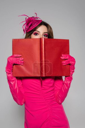young and brunette woman in magenta color gloves and hat with feather covering face with book isolated on grey 