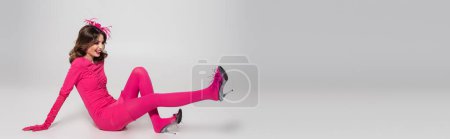 full length of happy young woman in hat and magenta color dress having fun on grey background, banner 