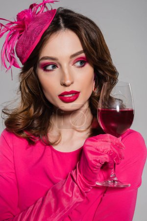 portrait of young woman in elegant hat and magenta color dress holding glass with alcohol drink isolated on grey 