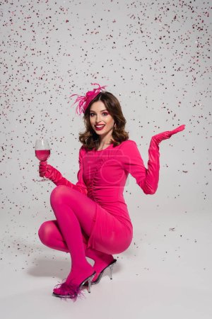 Téléchargez les photos : Full length of smiling woman in dress holding glass with magenta color drink on grey with falling confetti - en image libre de droit