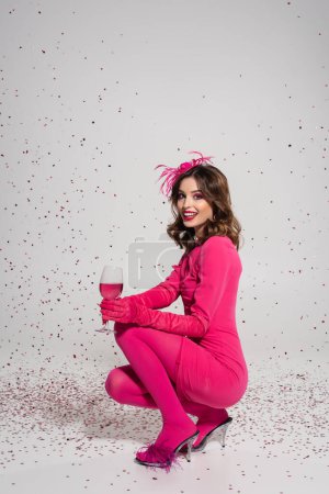full length of cheerful woman in dress holding glass with magenta color alcohol drink on grey with falling confetti 