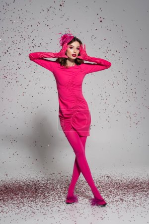 full length of brunette woman in elegant hat and magenta color dress posing on grey with falling confetti 