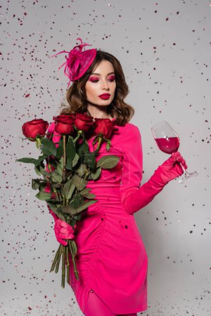 brunette woman in elegant hat and magenta color dress holding roses and glass of wine on grey with falling confetti 