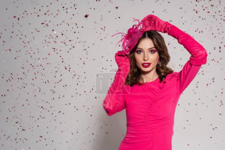 young brunette woman in magenta color dress adjusting elegant hat on grey with falling confetti 