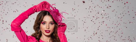 Téléchargez les photos : Young brunette woman in magenta color gloves adjusting elegant hat near falling confetti on grey, banner - en image libre de droit