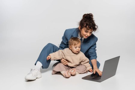 curly businesswoman in suit sitting with toddler daughter and using laptop on grey 